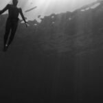 Water Activities - Black and white of from below of scuba diver swimming in clear water in swimsuit under rays of light