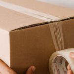 Accommodations - Crop anonymous male using tape to seal packed cardboard box against white wall while moving into new place