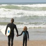 Beach Guides - A Man Carrying a Surfboard and a Child Walking on Beach Shore