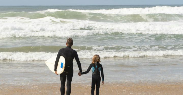 Beach Guides - A Man Carrying a Surfboard and a Child Walking on Beach Shore